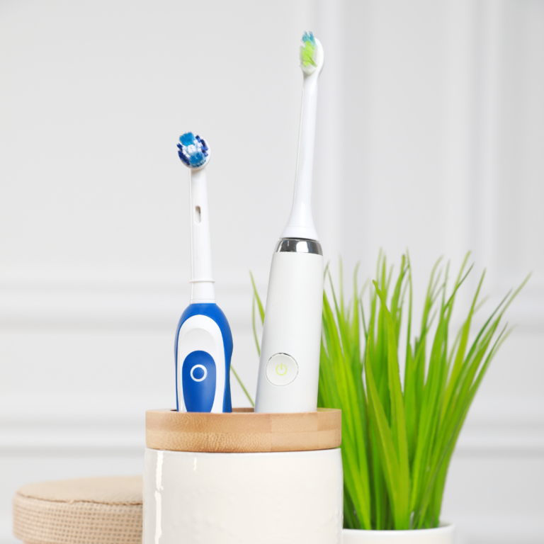 Image of two electric toothbrushes neatly placed in a cream and tan decorative vase, with a small leafy plant in the background.